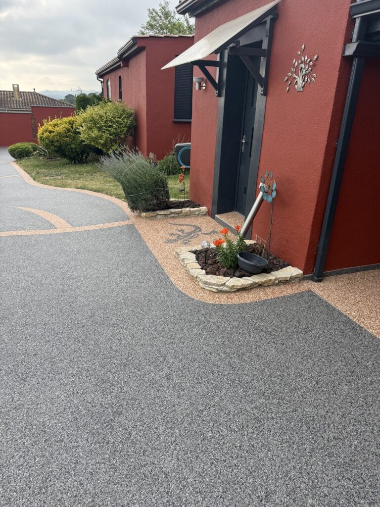 Maison rouge avec allée en moquette de pierre grise et beige, parterre fleuri et porte d'entrée moderne noire.
