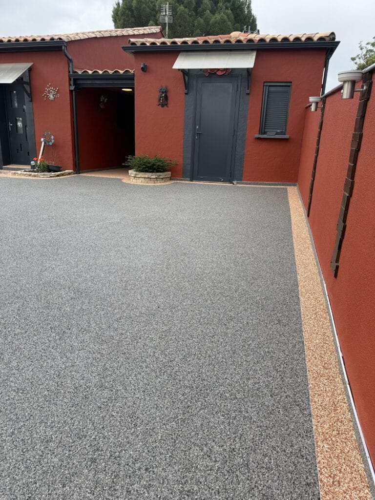 Revêtement en moquette de pierre grise et beige devant une maison rouge moderne aux menuiseries gris anthracite.