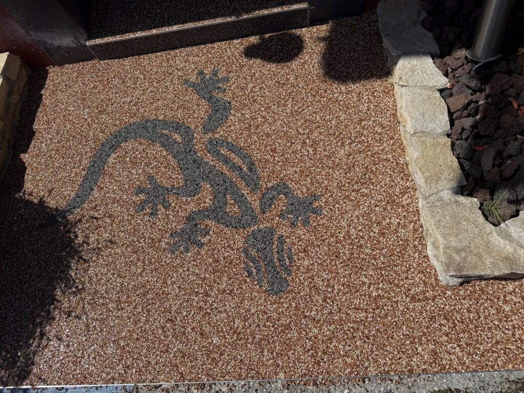 Motif de lézard noir en gravier résiné sur un sol décoratif brun et ocre avec bordures en pierre naturelle.