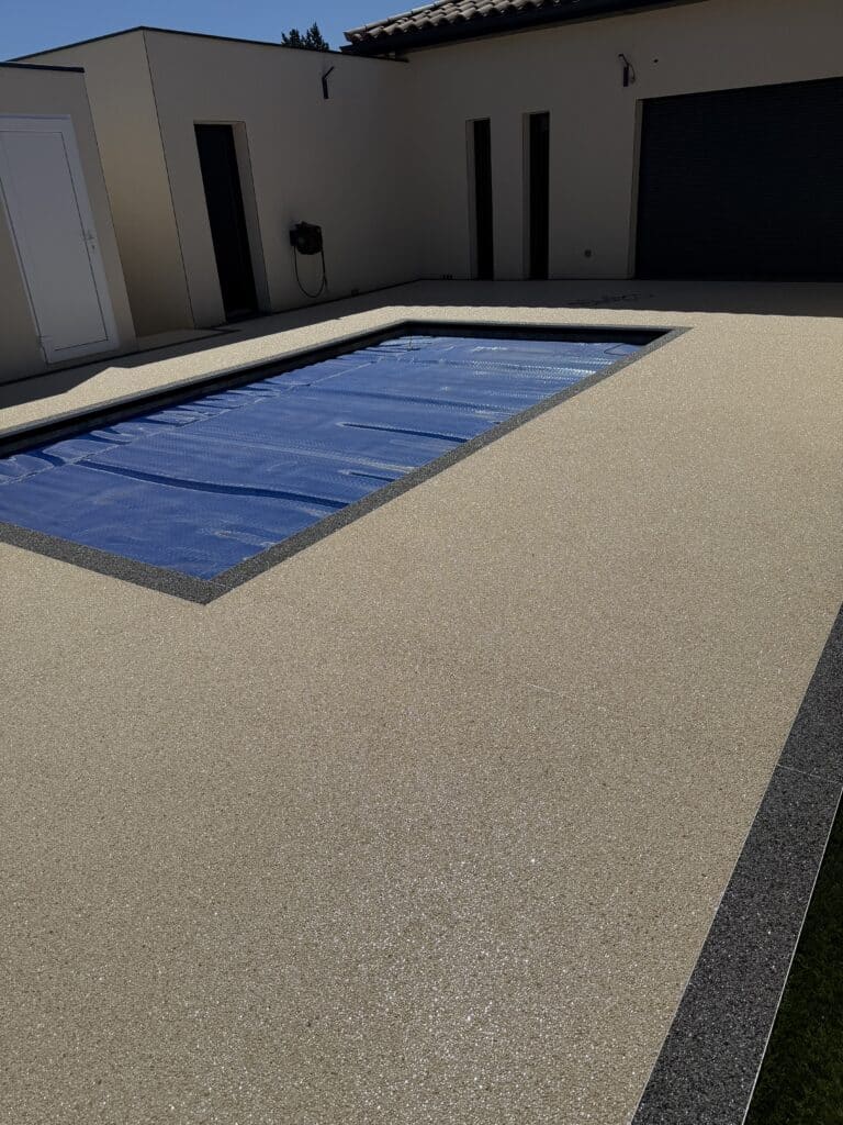 Piscine avec bâche bleue et terrasse en résine beige devant une maison moderne sous le soleil.